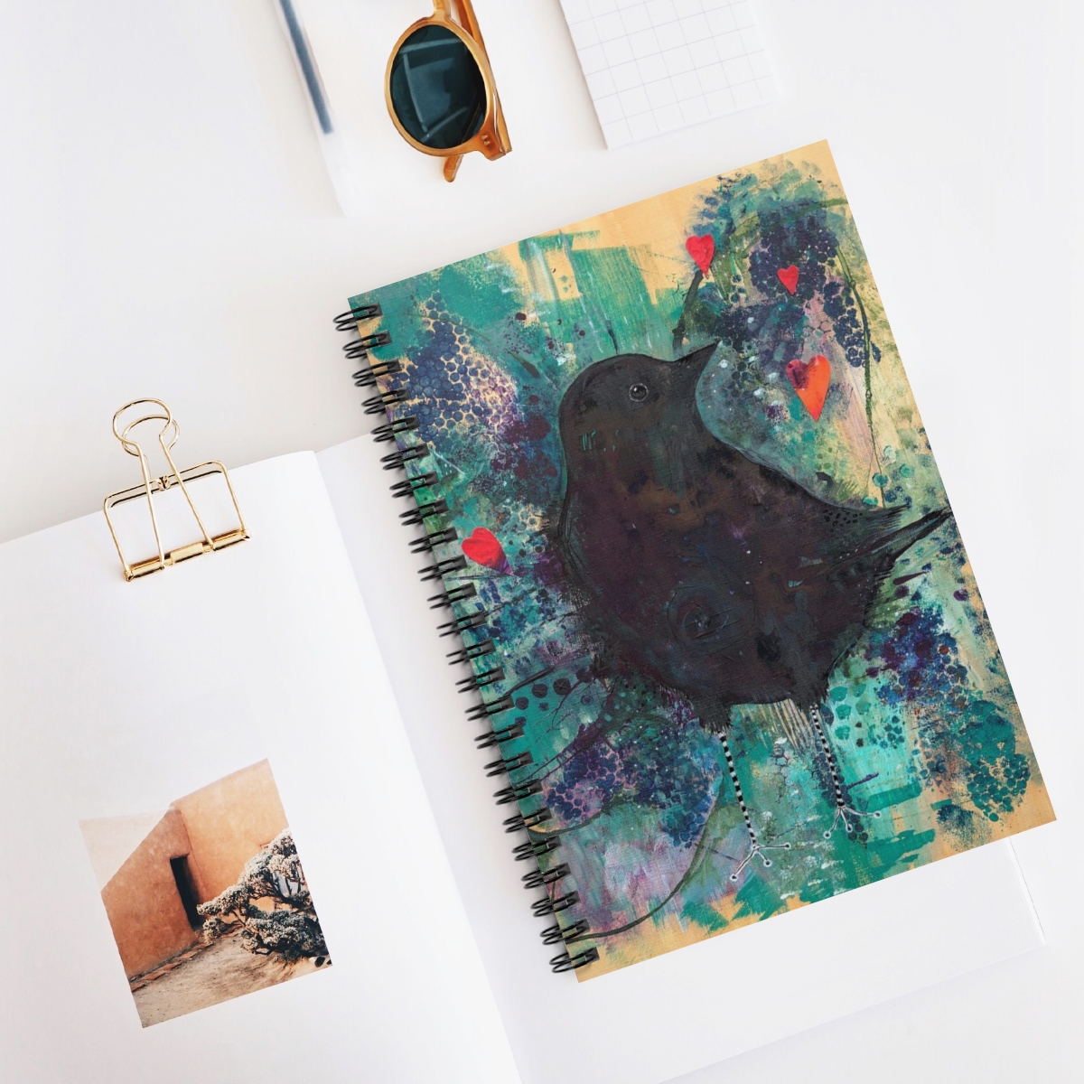 Special crow journal - this picture shows the crow notebook in context. The image on the notebook is a crow emerging from turquoise and purple toned and textured background. 3 red hearts float above him.