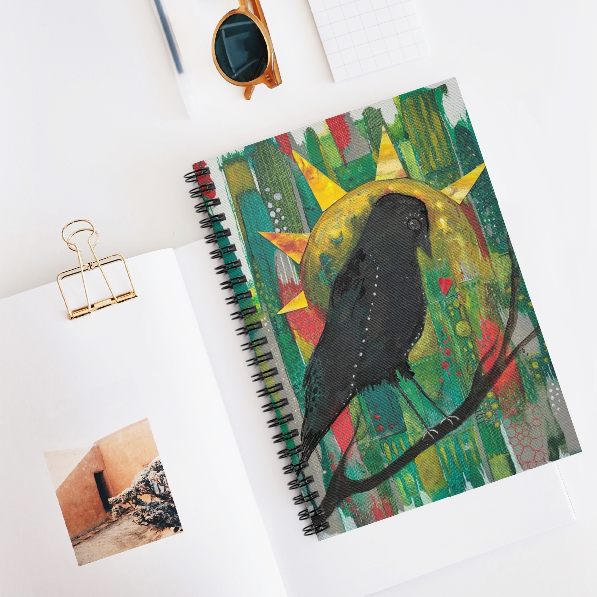 Special crow journal in context. Black crow sits on a branch surveying her patchwork kingdom of reds, greens and grey. Yellow sun behind her looks like her crown.