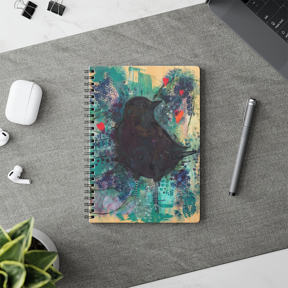 Special crow notebook - this picture shows the crow notebook in context next to a pencil. The image on the notebook is a crow emerging from turquoise and purple toned and textured background. 3 red hearts float above him.