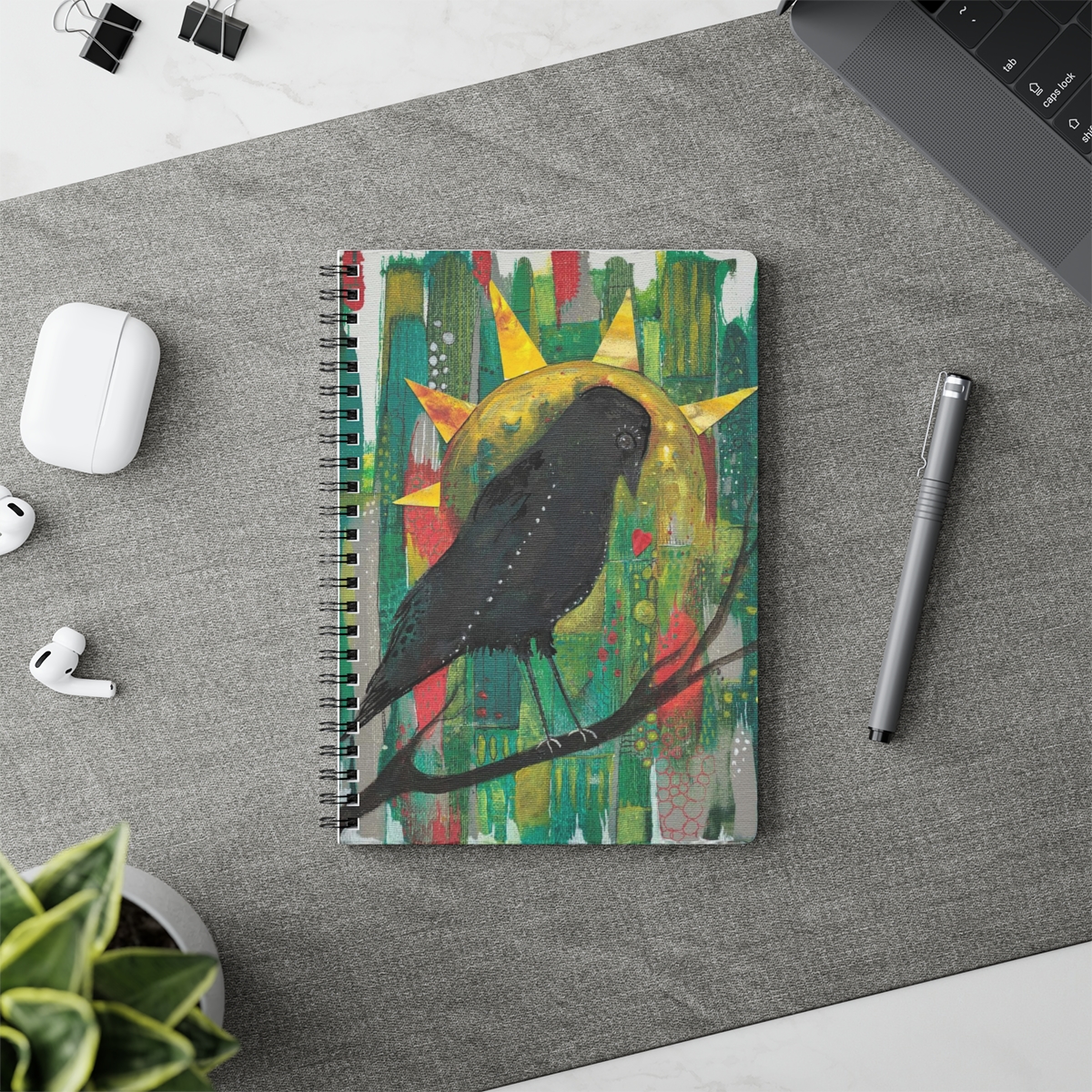 Special crow notebook in context. Black crow sits on a branch surveying her patchwork kingdom of reds, greens and grey. Yellow sun behind her looks like her crown.