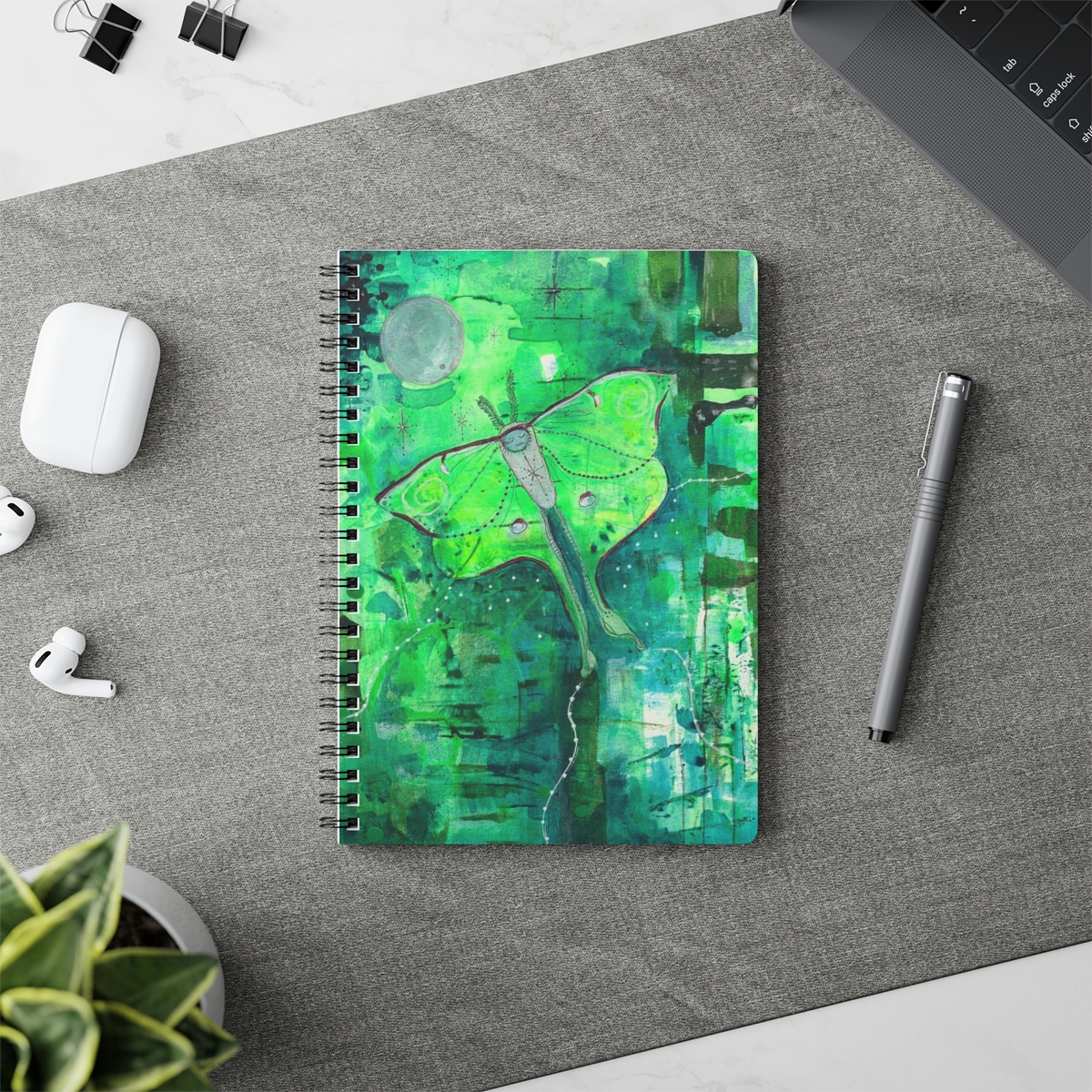 Special luna moth notebook in context. Luna moth is a whimsical painting. The moth flies against a background of striking greens towards the moon.