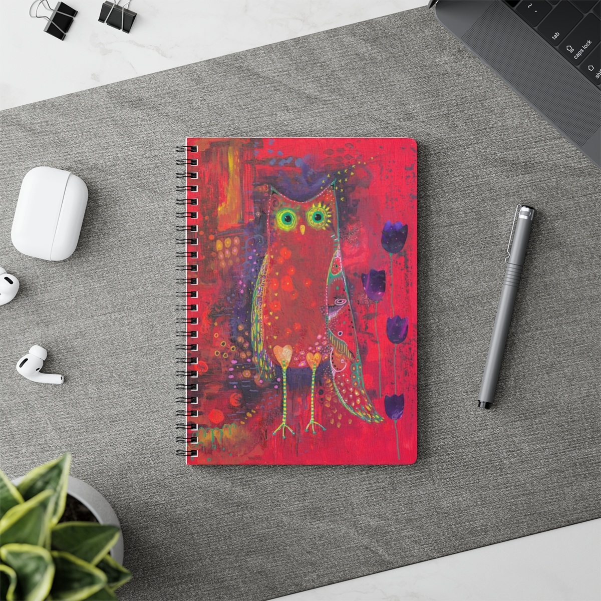 Owl notebook in context - Ruby Owl is a whimsical owl painted in hues of red and purple with yellow and green pattern detail. Purple tulips grow beside her.