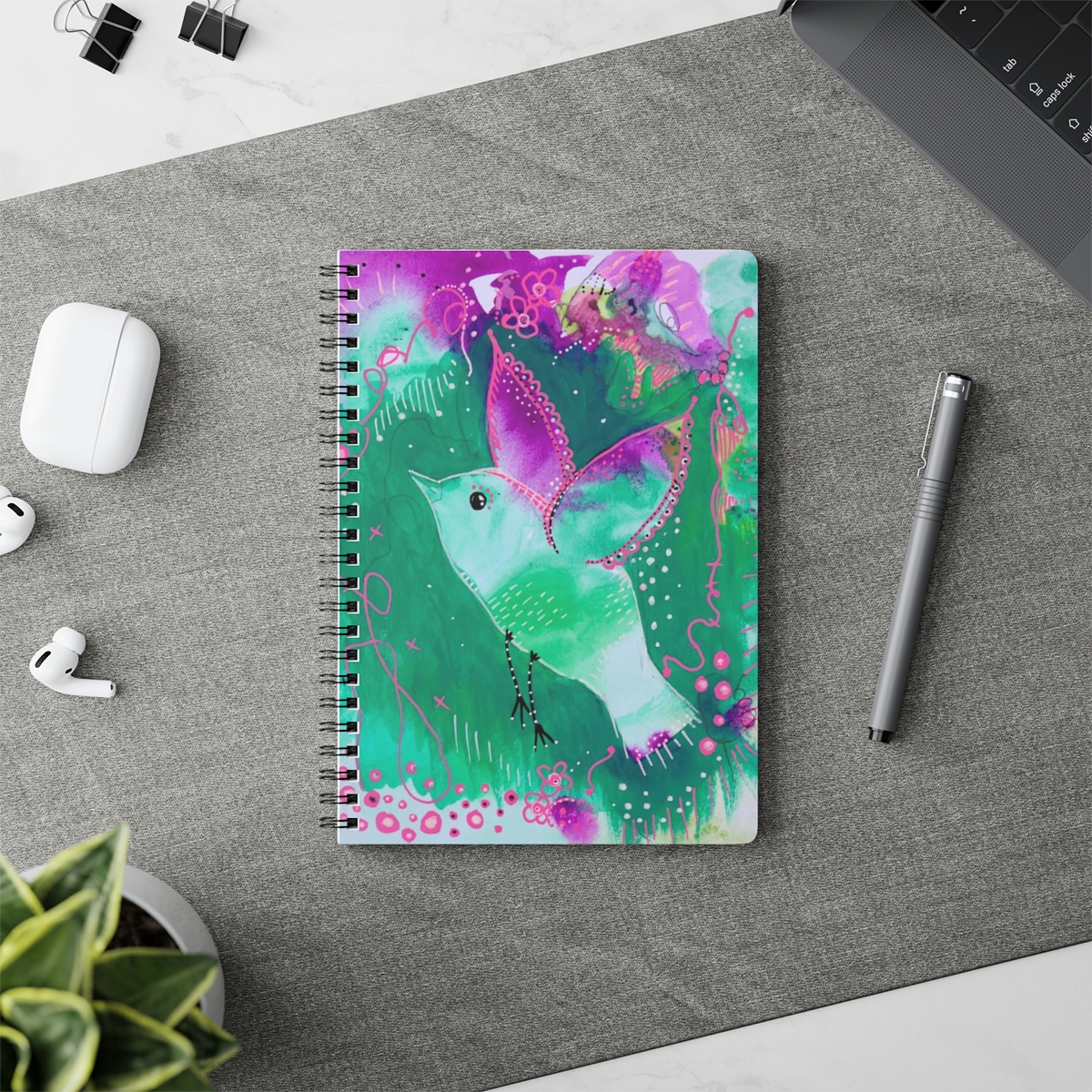 Whimsical bird notebook in context. Bird flying is created from green and violet tones that match her background.