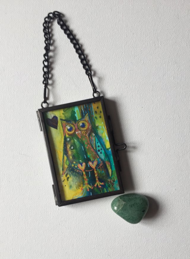 Special miniature owl - whimsical owl painted with greens, yellows and blues on an abstract background inside a glass hanging frame with crystal next to it to show size.