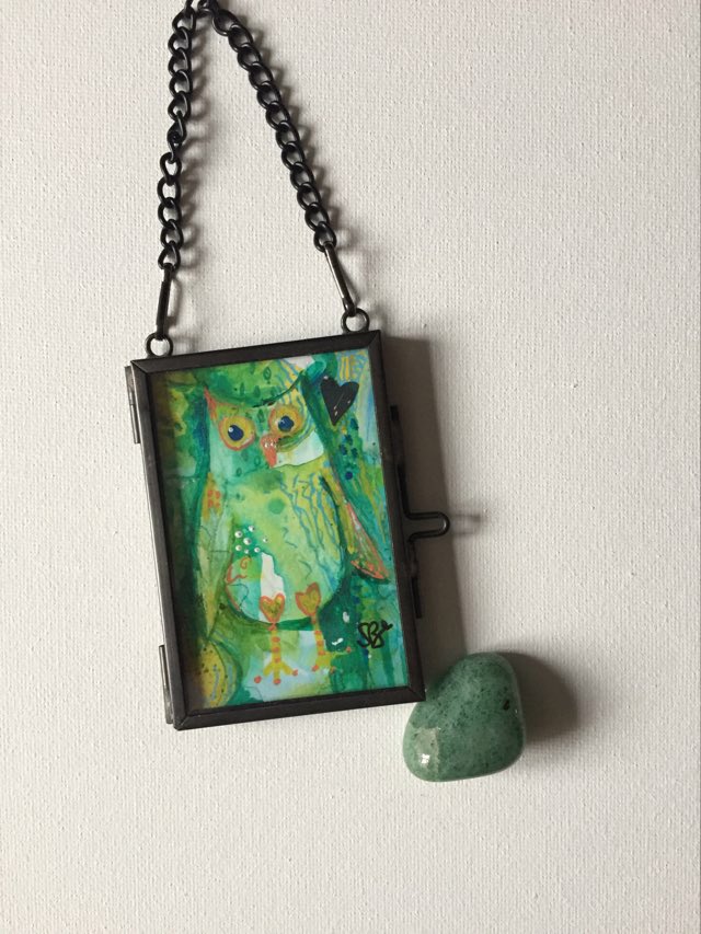 Special miniature owl painting - whimsical owl painted with greens, yellows and blues on an abstract background inside a glass hanging frame with crystal next to it to show size.