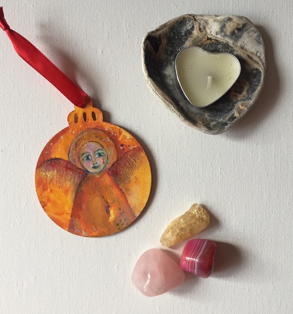 Hand-painted wooden bauble featuring an ethereal angel with a serene face, surrounded by yellow, orange and gold tones with hints of red wings. The ornament has a round shape with a decorative top, and the angel symbolises joy