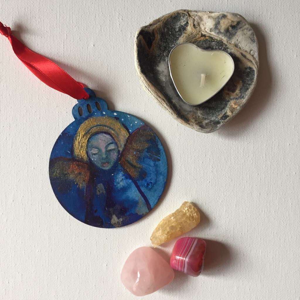 Hand-painted wooden bauble featuring an ethereal angel with a serene face, surrounded by blue and gold tones with hints of red wings. The ornament has a round shape with a decorative top, and the angel symbolises peace