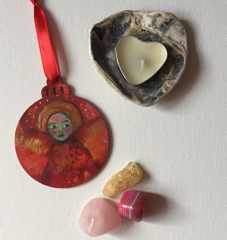 Hand-painted wooden bauble featuring an ethereal angel with a serene face, surrounded by red and gold tones with hints of red wings. The ornament has a round shape with a decorative top, and the angel symbolises love.
