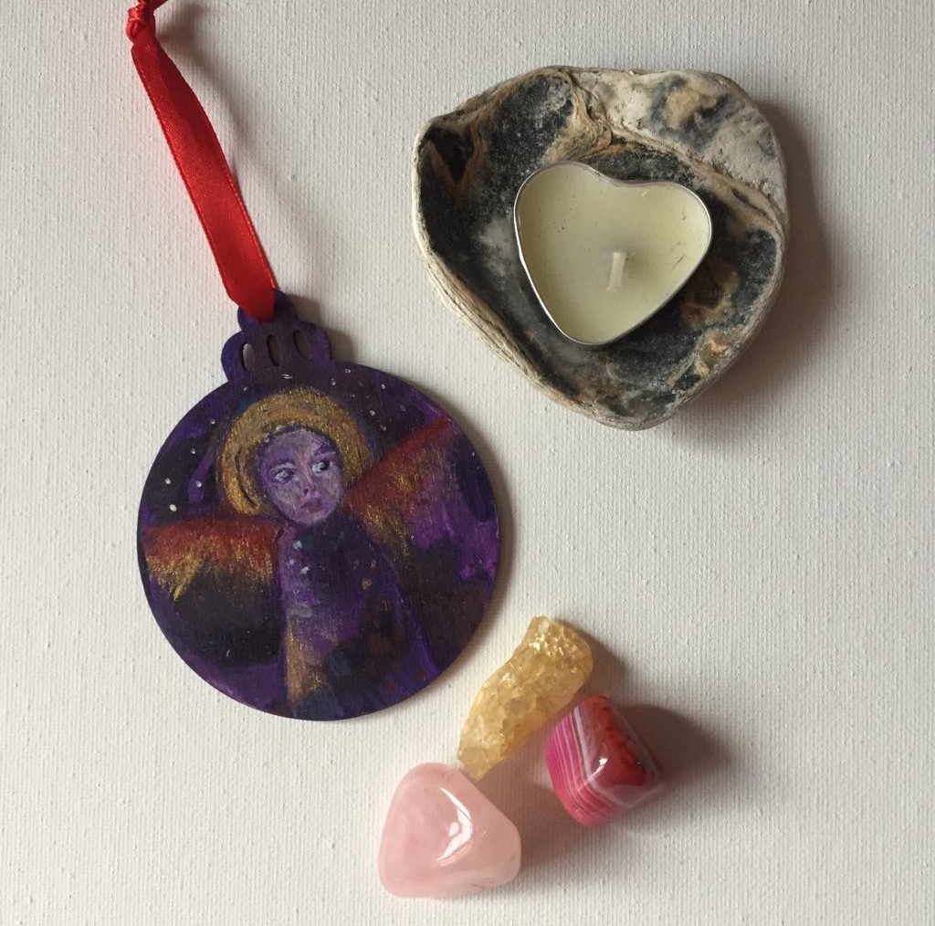 Hand-painted wooden bauble featuring an ethereal angel with a serene face, surrounded by purple and gold tones with hints of red wings. The ornament has a round shape with a decorative top, and the angel symbolises magic