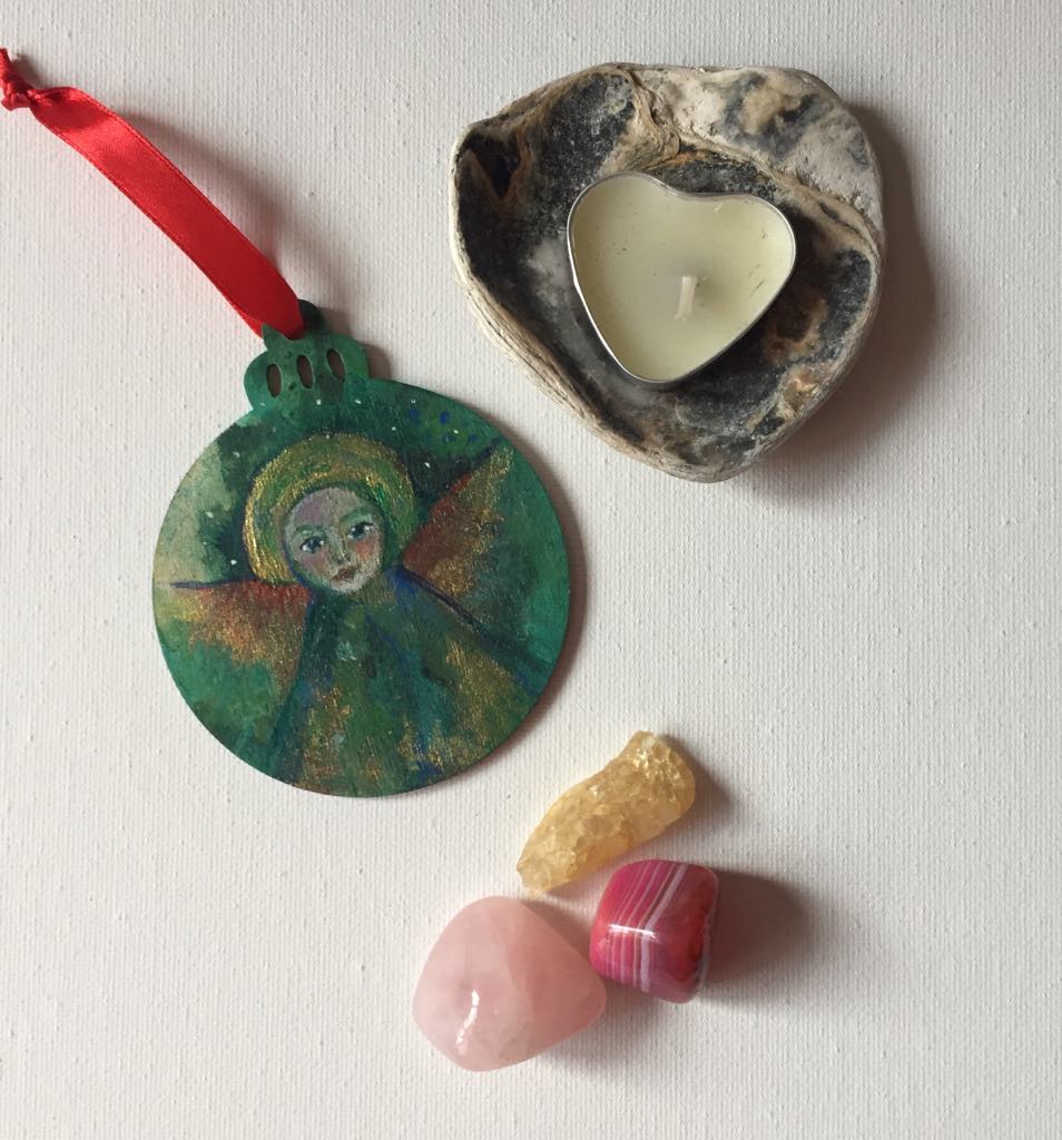 Hand-painted wooden bauble featuring an ethereal angel with a serene face, surrounded by green and gold tones with hints of red wings. The ornament has a round shape with a decorative top, and the angel symbolises Serenity.